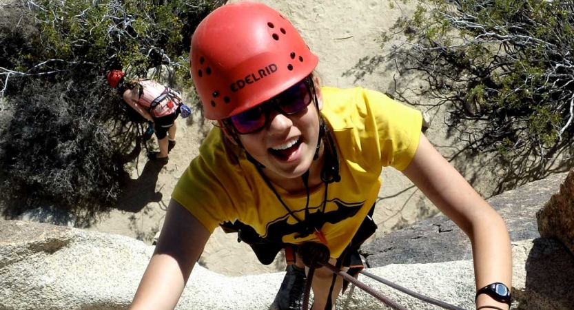 From above, a person wearing safety gear smiles as they climb towards the camera. 